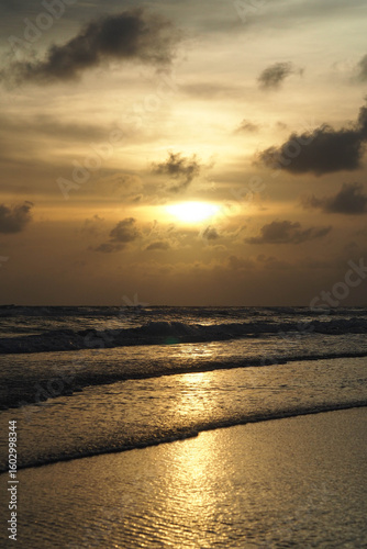 A beautiful sunset over the ocean with the sun's reflection on the waves and sand, under a cloudy sky.