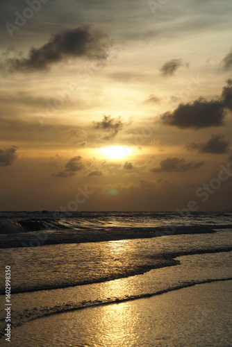 A beautiful sunset over the ocean with the sun's reflection on the waves and wet sand, under a cloudy sky.