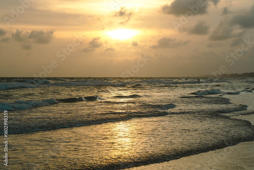A dramatic sunset over the ocean with the sun's reflection on the waves and wet sand, under a cloudy sky.