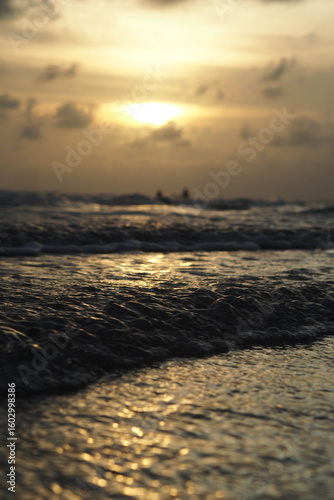 A golden sunset over the ocean with the sun's reflection on the waves, under a cloudy sky, and distant figures silhouetted against the horizon.