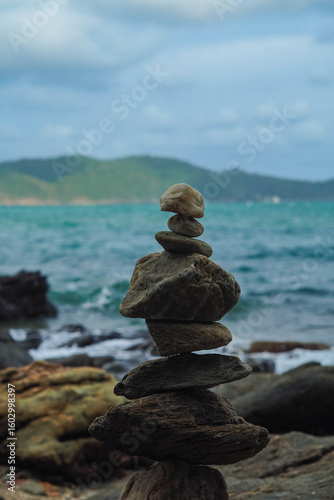 A stack of balanced rocks stands on a shore, with the ocean and distant mountains visible in the background.