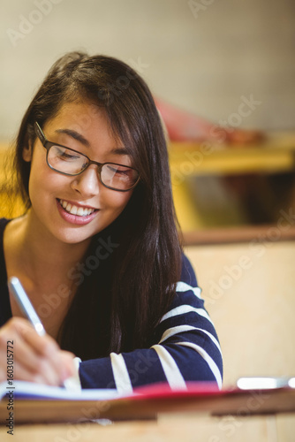 Asian woman sitting at desk in study room wearing glasses writing in notebook with silver pen