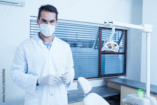 Male dentist wearing mask standing in clinic beside dental chair under overhead lamp, copy space