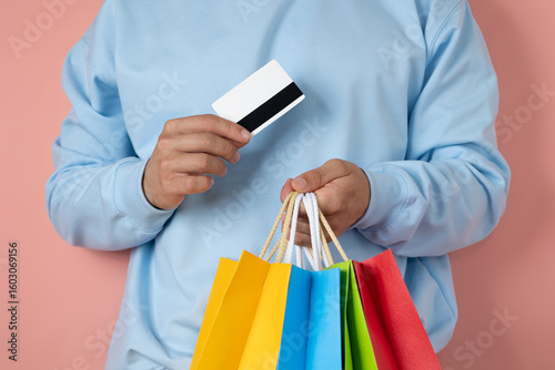 Close up of unrecognizable man in blue sweatshirt holding bank card and multi colored shopping bags on pink background