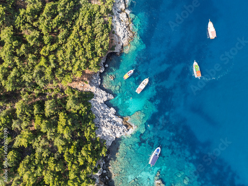 Fototapeta Naklejka Na Ścianę i Meble -  Aerial view of turquoise sea with boats near rocky coast and forest in Alanya, Turkey. Summer drone footage of scenic Mediterranean shoreline and clear water. Summer holiday in Alanya.