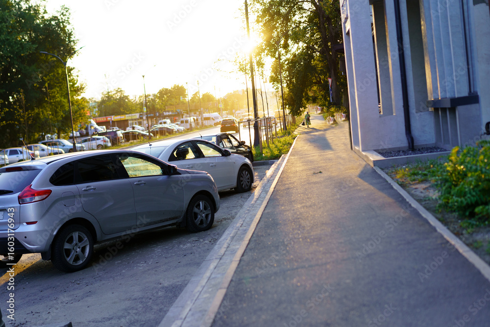 Evening sunlight illuminates parked cars along a quiet street beside a path in a residential area Evening sunlight illuminates parked cars along a quiet street beside a path in a residential area
