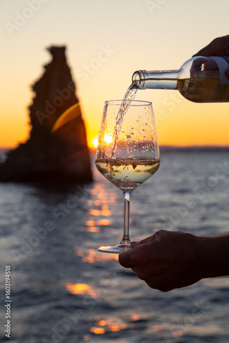 A person pours a glass of Albariño wine with the tower of San Sadurniño in the background. Silhouette at sunset.
