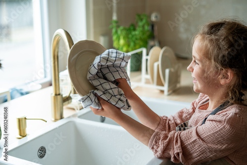 Little girl wipes clean washed plates with a towel. Child helps mother in the kitchen. Housework.
