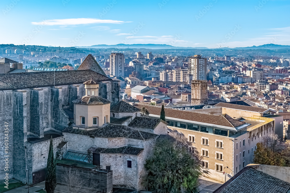 Obraz premium Panoramic view of Girona with Monastery of Sant Domenec, now part of the University of Girona. Historic stone buildings with terracotta roofs contrast with the modern cityscape and mountains