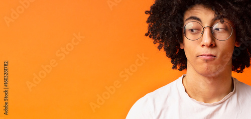 Black millennial man with expression of suspicion his face, looking skeptically aside at empty space, orange panorama background