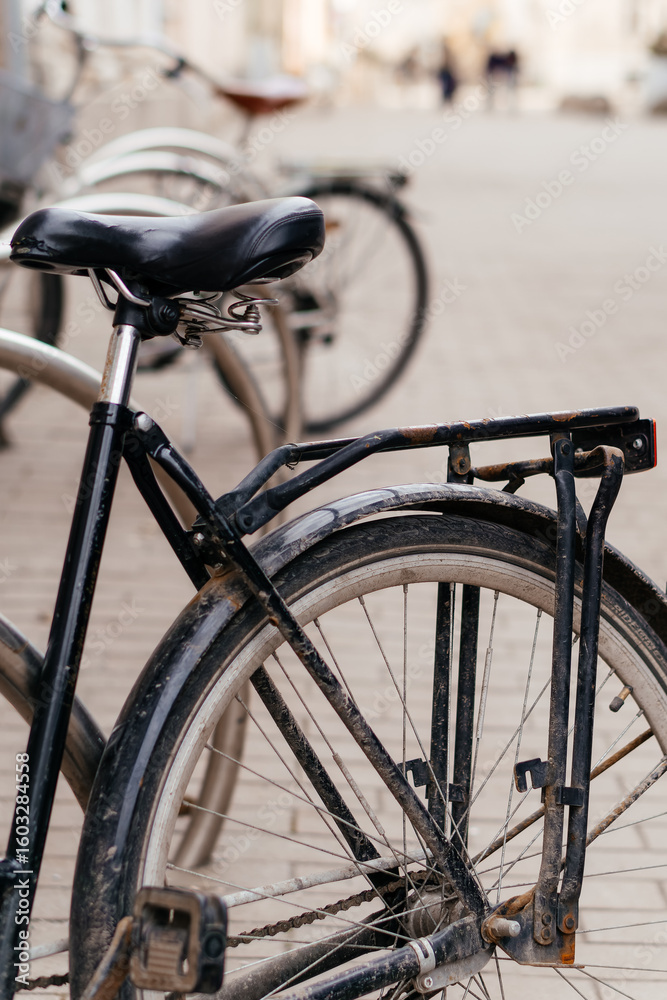 A close-up of an old, black bicycle with a worn-out frame and dusty wheels, parked among other bikes in an urban setting A close-up of an old, black bicycle with a worn-out frame and dusty wheels, parked among other bikes in an urban setting