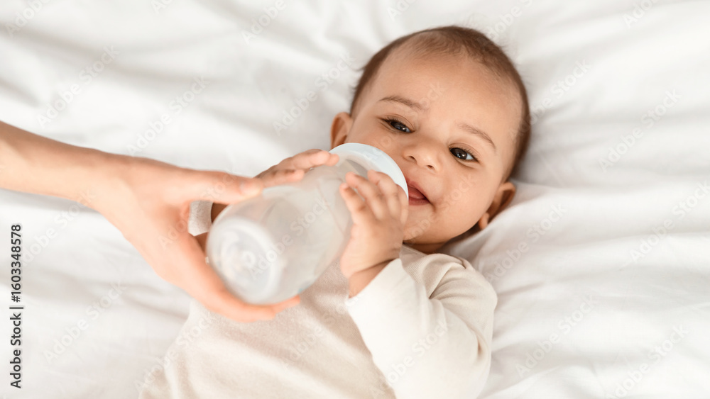 Obraz premium Nursery And Childcare Concept. Portrait of cute little black kid drinking milk, mother holding baby bottle in hands, feeding her child who lying on white blanket on the bed in the bedroom at home