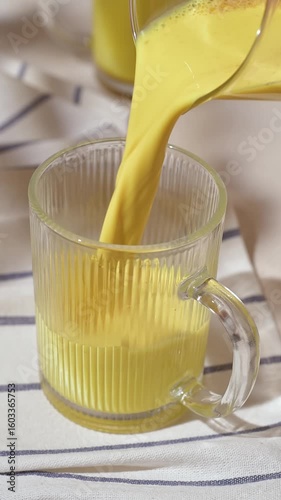 Golden latte pours into clear ribbed mug, creating cozy healthy moment with warm yellow color on striped morning textile.