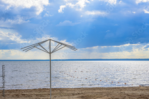 Empty beach with metal umbrella frame on sandy shore, cloudy sky over calm sea, peaceful coastal scene with overcast weather, deserted seascape with soft light and dramatic cloudscape