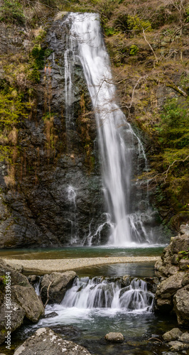 A tall waterfall in Minoh National Park, Osaka, Japan. The image has a long exposure, highlighting the water's smooth flow. Rocks and lush vegetation frame the scene, evoking a sense of tranquility.