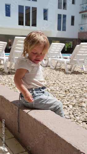 A blond toddler's day evolves from a distressed outcry on a sunny patio to quiet play and then peaceful contemplation by a resort pool.