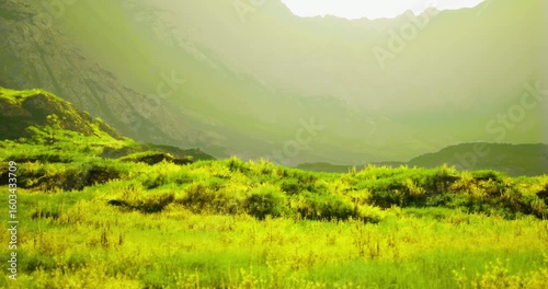 A lush green landscape features rolling hills and towering mountains bathed in bright sunlight. The scenery highlights the beauty of nature during daytime, evoking tranquility.