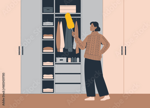 A young woman organizes her closet with a dusting brush, representing the importance of cleanliness and tidiness in living spaces, highlighting her proactive approach to home care.