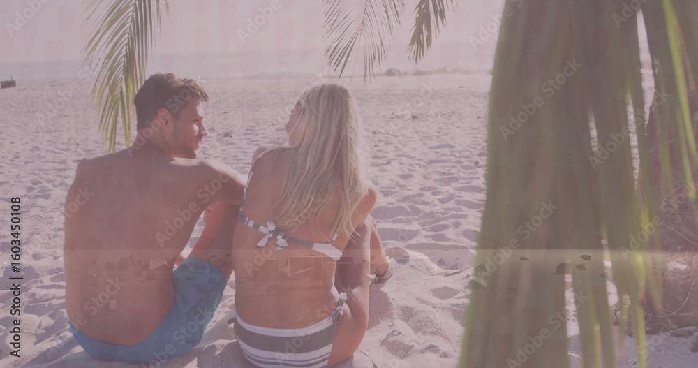 Fototapeta premium Relaxing adult couple wearing swimwear sitting on beach at sunset, with palm fronds and ocean waves
