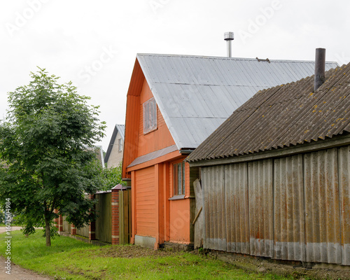 Fototapeta Naklejka Na Ścianę i Meble -  Bright orange wooden house with metal roof stands beside a weathered shed in a quiet rural village neighborhood with greenery.