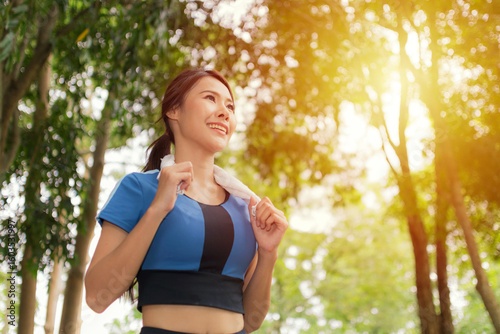 Smiling Asian woman with a towel around her neck, jogging in a park, embodying joy and a healthy, active lifestyle.