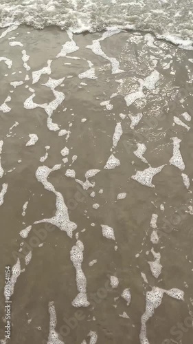 A calm wave reaches the sandy beach, leaving behind scattered bubbles on the smooth surface