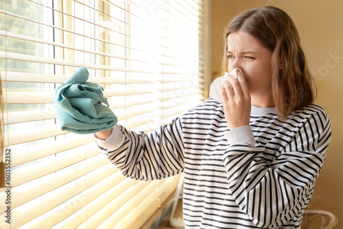 Fotografie Young woman suffering from dust allergy while cleaning window blinds