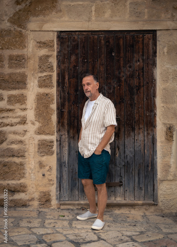 Senior man in summer outfit standing by rustic wooden door.