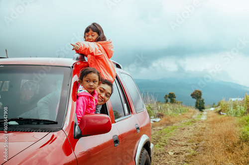 Happy Asian mother with her two little daughters peeking out from car window, enjoying nature scenery on their road trip adventure through the mountain track