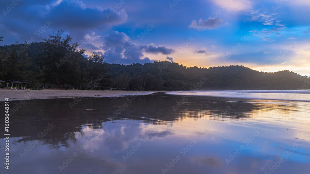 Obraz premium Evening after sunset on a tropical island. The clouds in the blue sky are highlighted in pink and gold. Silhouettes of mountains. Reflection on the smooth shiny surface of the ocean. Malaysia. Borneo.