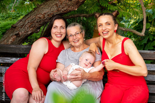 Three women from different generations pose happily on a park bench outdoors, with a baby in the grandmother's lap, surrounded by green trees and foliage