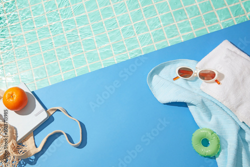 Bright summer setup with blue tiles, white towel, green float, and orange fruit near turquoise water, with soft space for product or design.