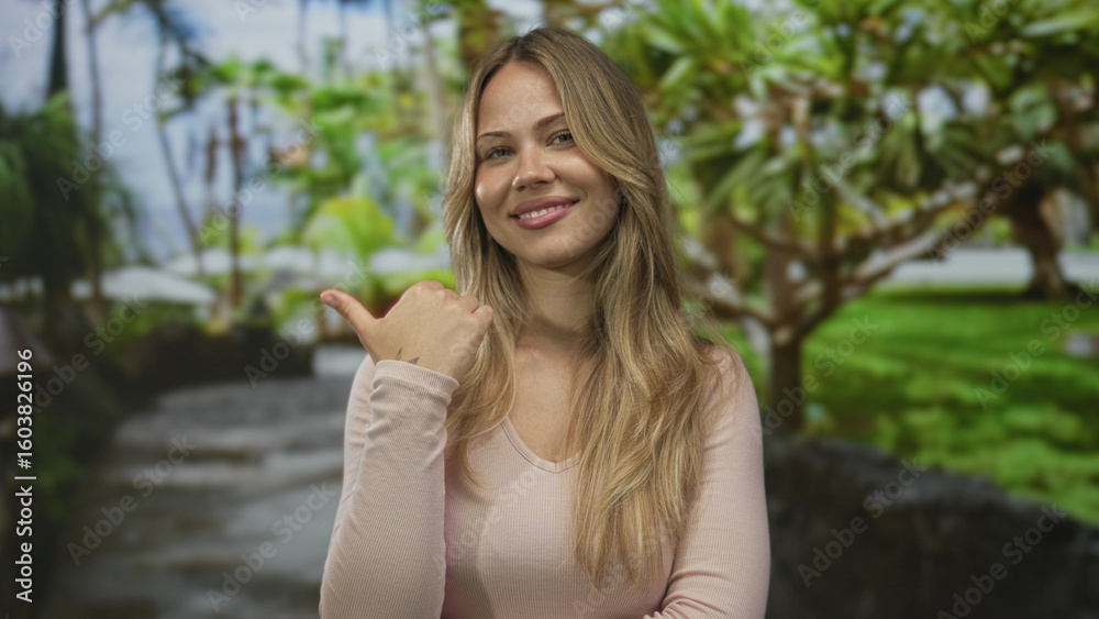 Naklejka premium Blonde woman pointing thumb to side on a sunny park walkway with tropical trees; direction confidence.