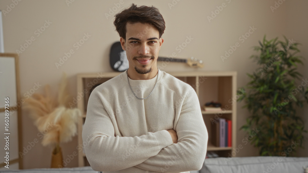 Fototapeta premium Young man with crossed arms standing confidently in a cozy living room, exuding a sense of calm and composure, surrounded by home decor elements in a modern apartment.