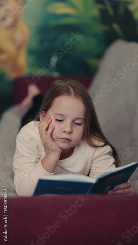 A young girl lies on a couch, engrossed in her book. The blurred background suggests a cozy and comfortable atmosphere, perfect for quiet reading.