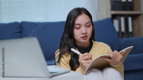 Young asian woman taking notes while studying on laptop at home