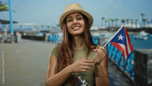 Tableau sur toile Woman smiling holds puerto rican flag at port under blue sky wearing hat with camera capturing her outdoor adventure in vibrant setting celebrating travel culture and patriotism