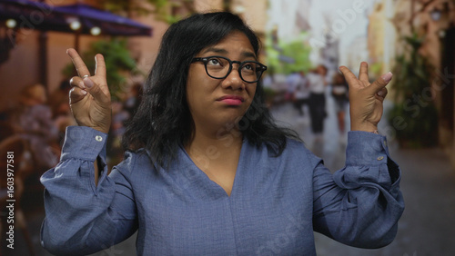 Photos Woman wearing glasses and a blue blouse making finger quotes gesture and rolling eyes on street; sarcasm skepticism