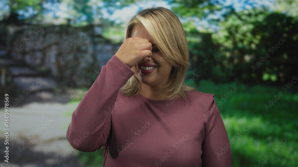 Woman wearing purple sweater grimaces while pinching her nose on sunlit park path with green grass; disgust. Woman wearing purple sweater grimaces while pinching her nose on sunlit park path with green grass; disgust.