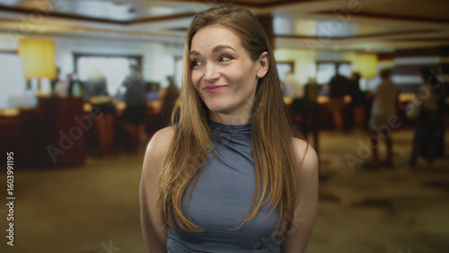 Young caucasian woman makes money gesture by pinching fingertips in hotel building lobby with a skeptical smile; greed.