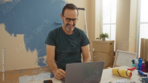 Man smiling in new living room surrounded by unpacked boxes and painting supplies, suggesting a fresh start in a cozy home setting.