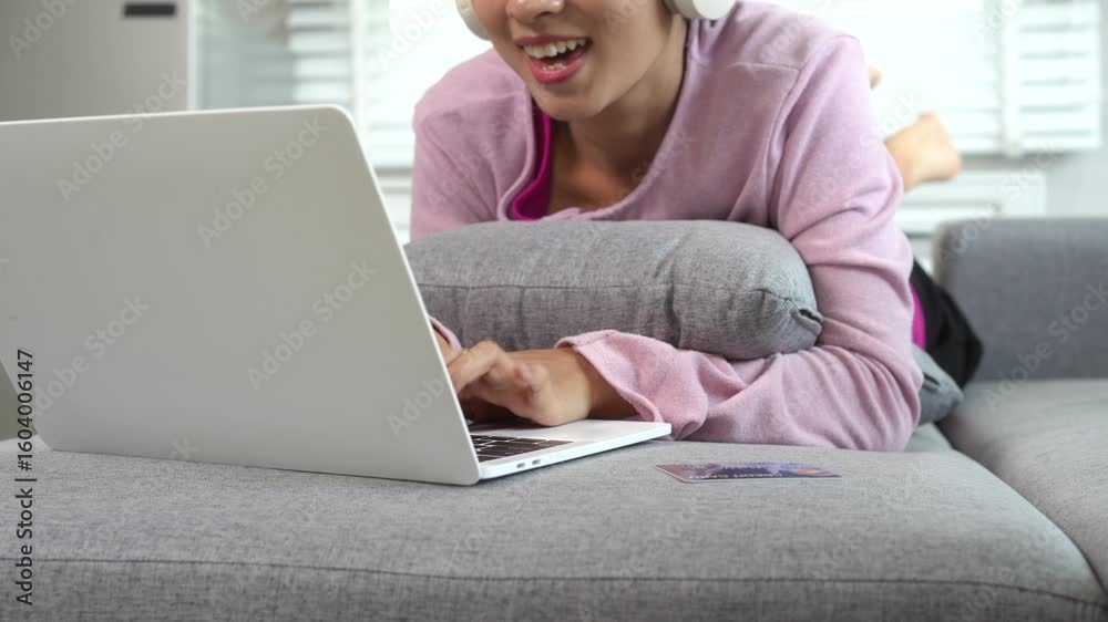 A young Asian housewife relaxes on the sofa, wearing headphones and smiling while shopping online. She holds credit card and uses her laptop to manage household purchases in cozy home environment. A young Asian housewife relaxes on the sofa, wearing headphones and smiling while shopping online. She holds credit card and uses her laptop to manage household purchases in cozy home environment.