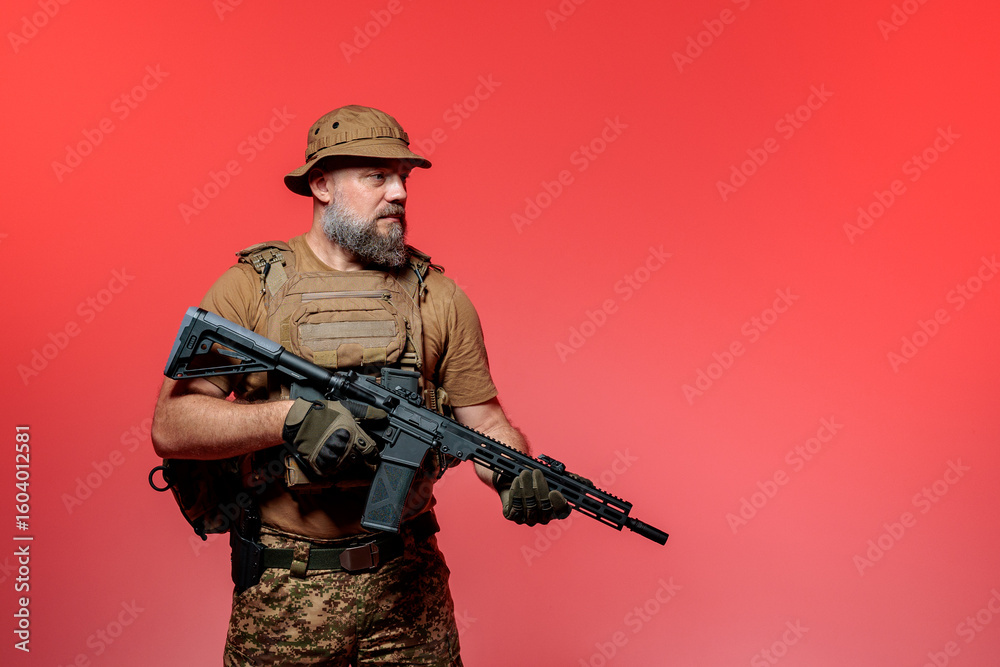 Fototapeta premium A soldier in military uniform holds a rifle AR 15 in his hands on red background in studio