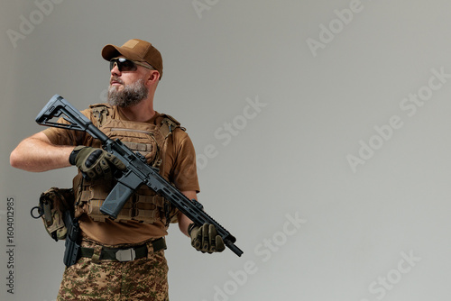 A soldier in military uniform holds a rifle AR 15 in his hands on grey background in studio