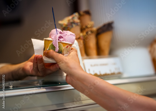 Close-up of a paper cup of purple gelato being handed over a counter, with blurred waffle cones in the background, capturing a friendly ice cream shop moment.