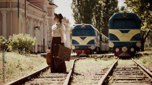Woman in vintage clothes standing on railroad with suitcase near two blue trains. Concept of historical travel, period fiction story, culture in transition, romantic narrative.