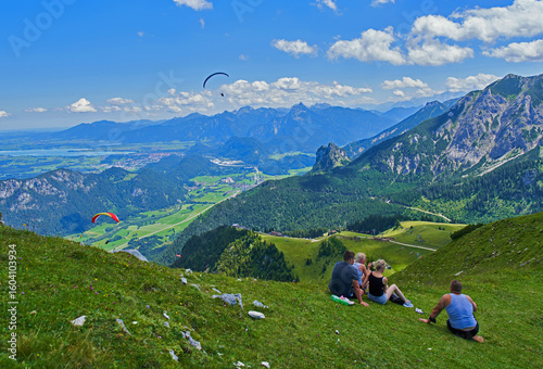 Pfronten, Germany - 28 July 2020: View of lush green slopes where people sit, contrasted against the distant rugged mountains and paragliders in the clear blue sky.