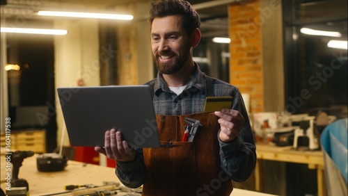 Bild auf Leinwand Cheerful Caucasian man wood master hold credit card typing laptop in workshop ma