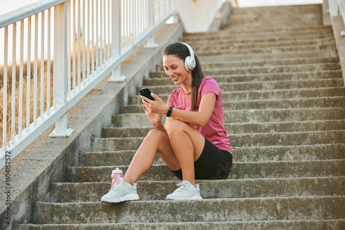 Photos A person is sitting on outdoor stairs, wearing headphones and smiling while looking at a smartphone