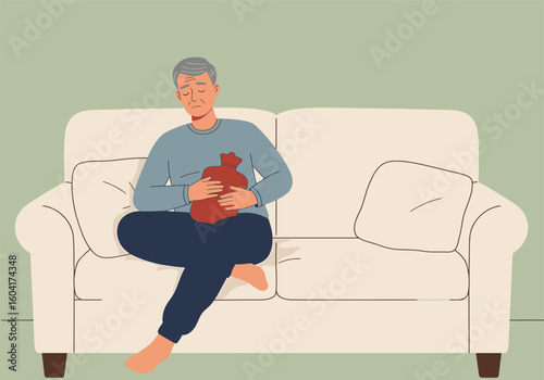 An elderly man with a stomach ache sits on a couch holding a red hot water bottle for pain relief.